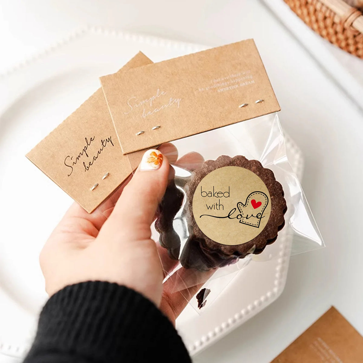 A hand holds a clear plastic bag containing kraft paper seal labels and a dark chocolate cookie decorated with a heart.