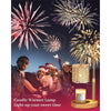 A golden candle warmer lamp with a glass jar sits on a wooden base, set against a background of fireworks.