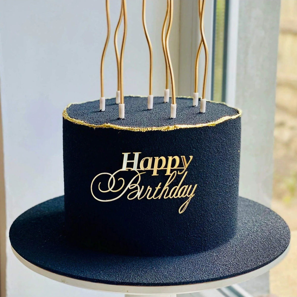 A dark navy blue birthday cake with gold lettering and thin acrylic toppers on a black cake board.