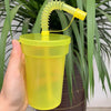 A hand holds a neon yellow reusable sipper cup with a straw and lid, set against a backdrop of green foliage.