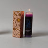A glass candle in a dark cylinder with purple wax is lit, next to a decorative box featuring zodiac symbols.