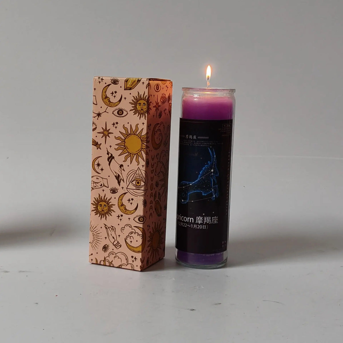 A glass candle in a dark cylinder with purple wax is lit, next to a decorative box featuring zodiac symbols.