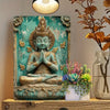 A bronze statue of a meditating deity with golden crown and hands in prayer set against a teal background.