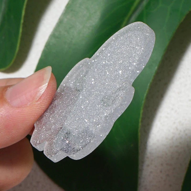 A hand holds a clear silver glitter acrylic charm against a green leaf background.