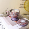 A glass candle in a patterned tin sits on a white book next to a sprig of greenery.