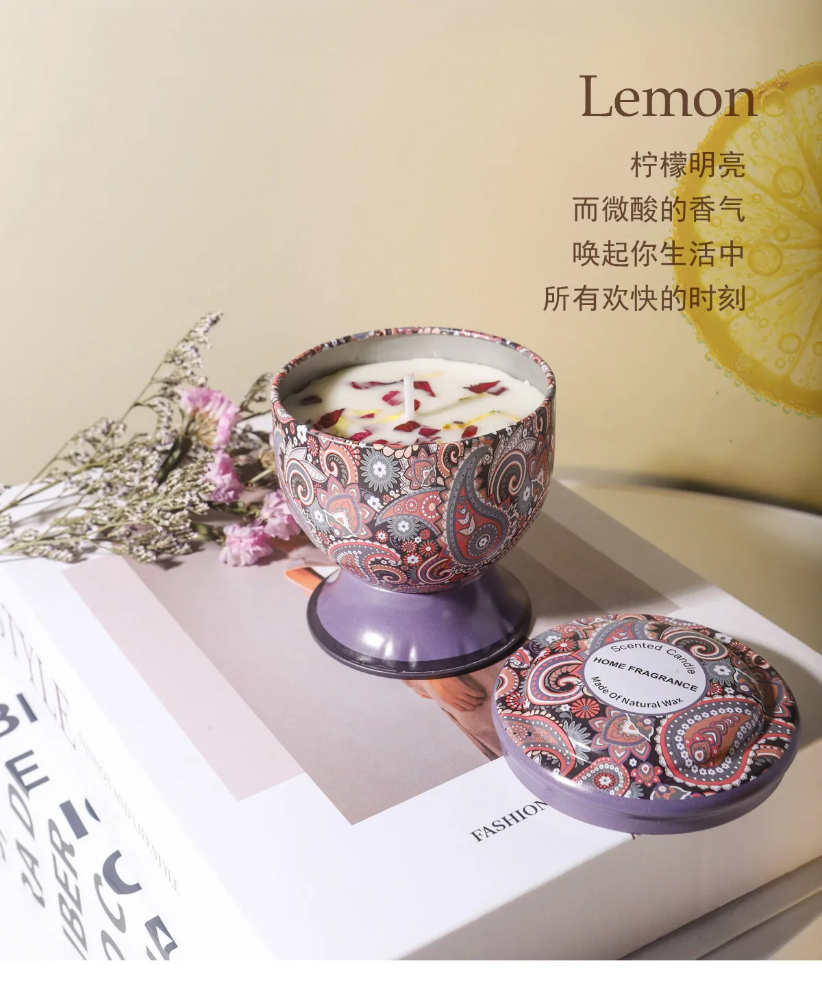 A glass candle in a patterned tin sits on a white book next to a sprig of greenery.