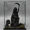 A black figurine of a gothic female grim reaper wearing a hood and holding a scythe, displayed in a glass box.
