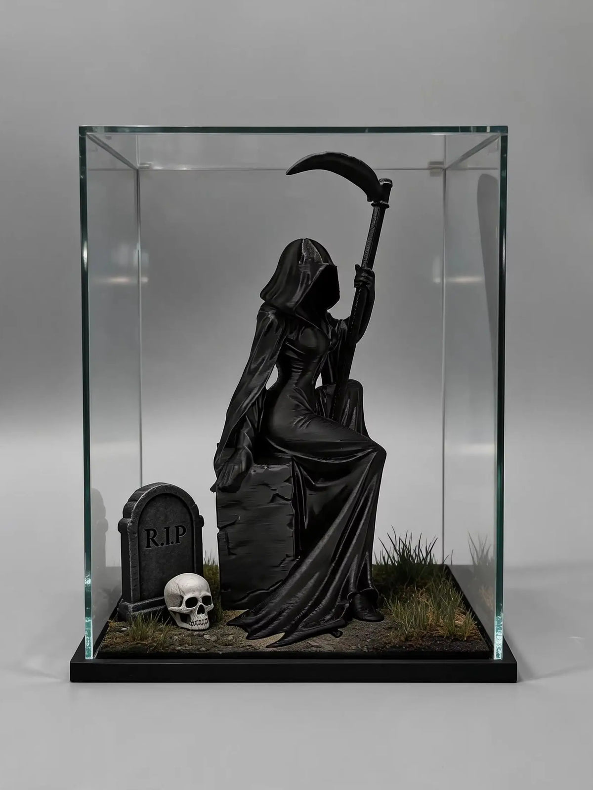 A black figurine of a gothic female grim reaper wearing a hood and holding a scythe, displayed in a glass box.