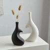 A modern heart-shaped ceramic vase in matte black and white sits on a white surface with dried ornamental grasses.