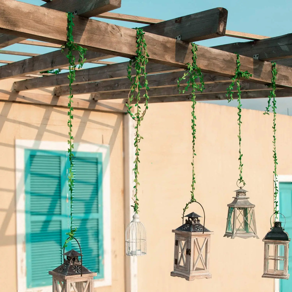 Artificial green climbing vine hanging from a wooden pergola structure against a light yellow wall.