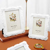 A white ornate photo frame with a floral design is displayed on a wooden surface next to a gold stand and a magnifying glass.