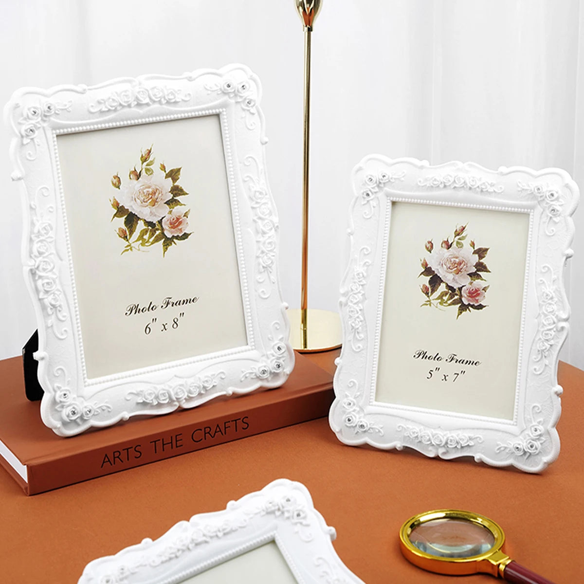 A white ornate photo frame with a floral design is displayed on a wooden surface next to a gold stand and a magnifying glass.