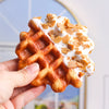 A hand holds a golden waffle topped with white icing and sliced almonds.