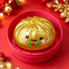 A golden squishy stress reliever shaped like a smiling snowman with pink cheeks and a green bow, resting in a pink container.