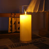 A tall glass candle with a flickering yellow flame is lit in a dimly lit room.