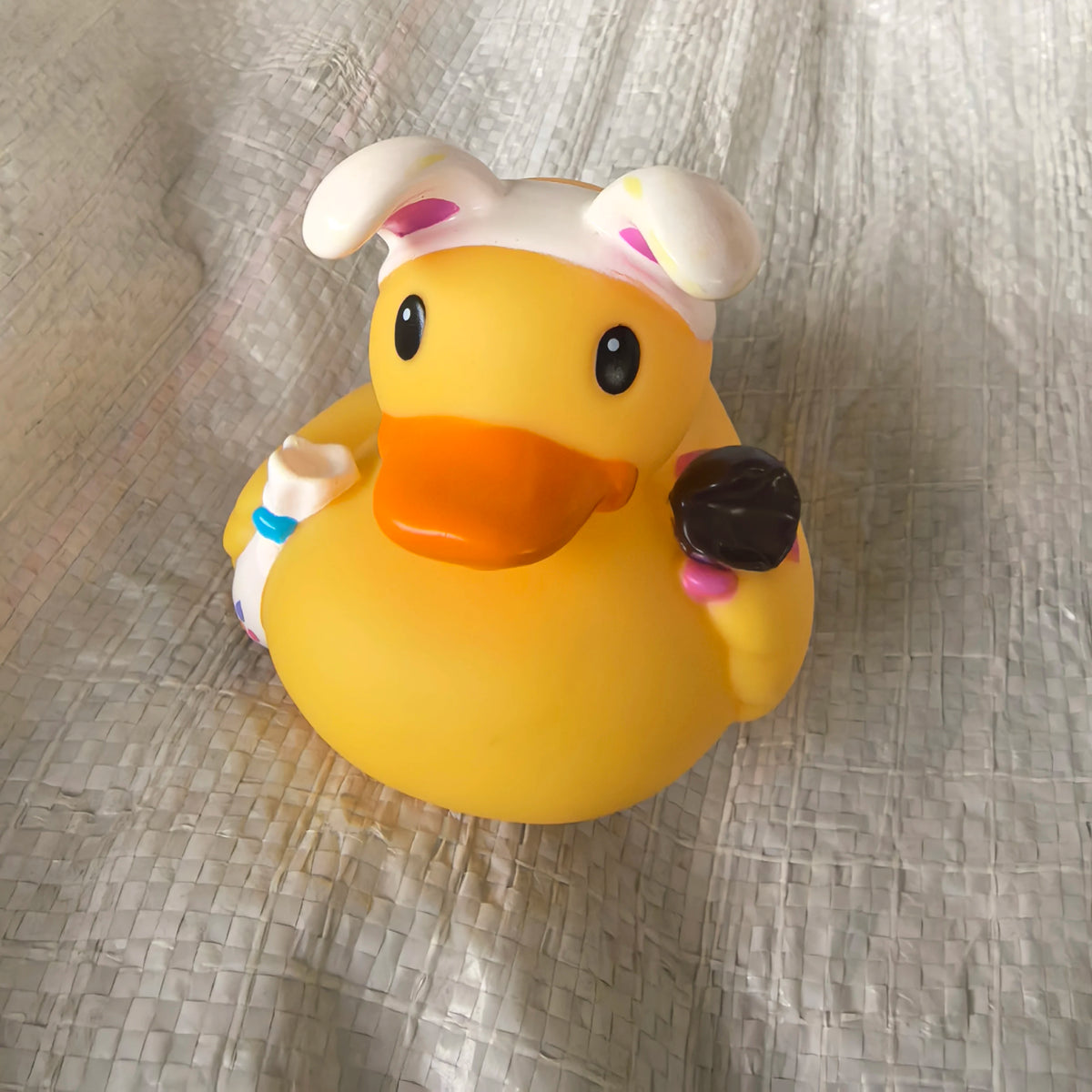 A yellow rubber duck bath toy with white and pink accents is shown on a wrinkled light-colored cloth.