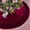 A deep red velvet Christmas tree skirt with a ruffled edge decorated with gold ornaments and green pine branches.