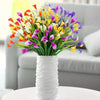 A vibrant bouquet of artificial calla lilies in shades of purple, yellow, and orange displayed in a white vase on a sofa.