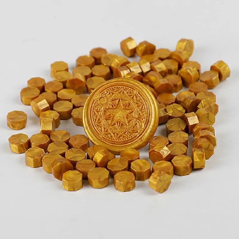A pile of small, round, gold-colored wax sealing beads and one larger circular bead with a star pattern.