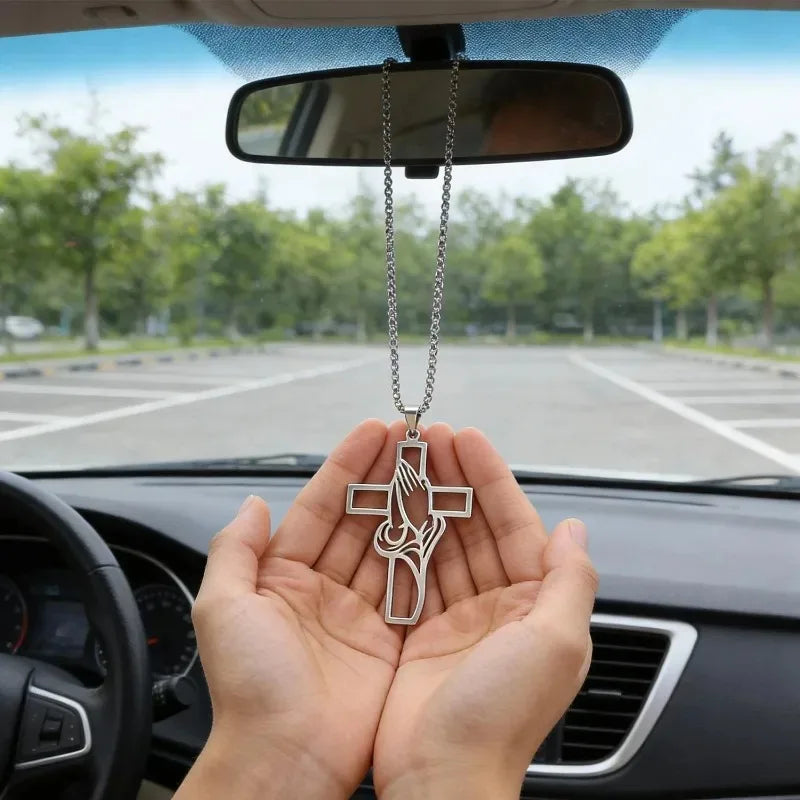 A hand holds a stainless steel praying hands cross pendant against the backdrop of a car mirror.