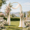 A gold metal arch backdrop stand set featuring white draping and floral arrangements in a garden setting.