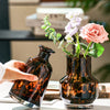 A hand holds a dark amber glass vase with leopard print patterns, next to another similar vase.