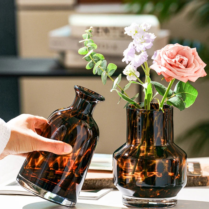 A hand holds a dark amber glass vase with leopard print patterns, next to another similar vase.