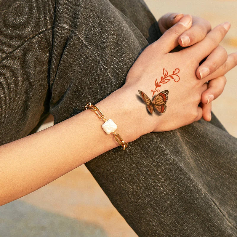 A hand wearing a gold bracelet and featuring a butterfly temporary body art sticker on the wrist against dark clothing.
