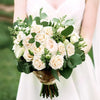 A bouquet of cream and blush roses with green foliage held by a person in a white dress.