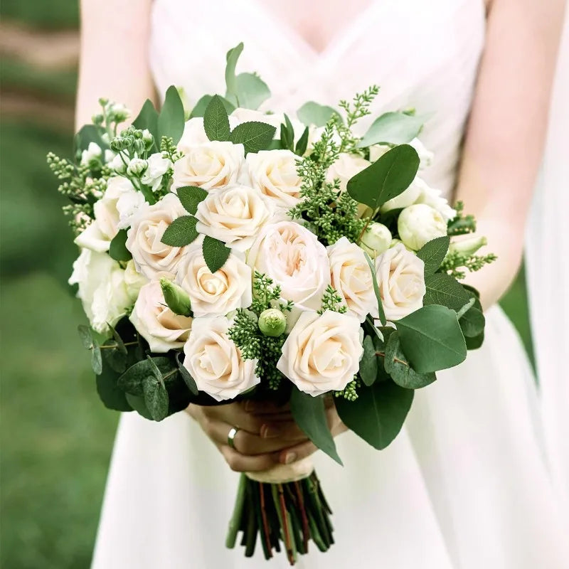 A bouquet of cream and blush roses with green foliage held by a person in a white dress.