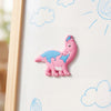 A pink dinosaur fridge magnet with blue spots is displayed on a white board with light blue clouds and sun rays.