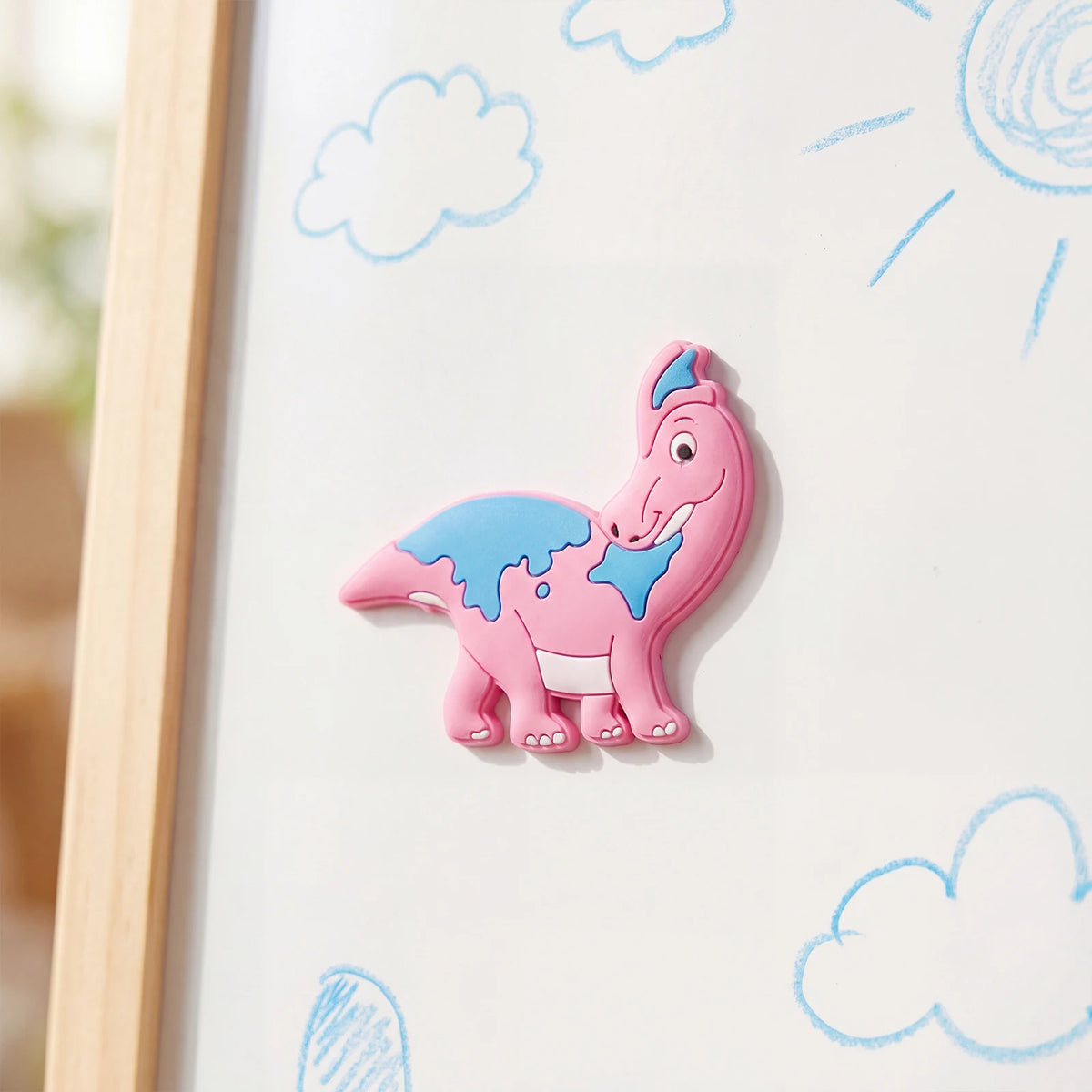 A pink dinosaur fridge magnet with blue spots is displayed on a white board with light blue clouds and sun rays.