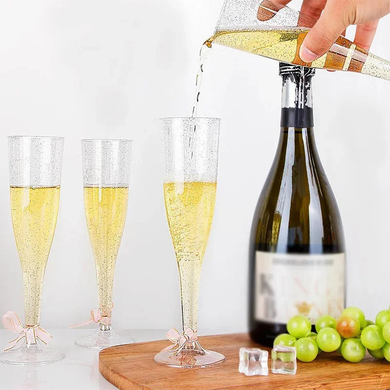 A hand pours sparkling yellow liquid into a glass champagne flute, with two empty flutes and green grapes nearby.