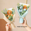 A hand holds a bouquet of handmade sunflower and daisy crochet flowers wrapped in light blue paper.