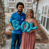 A man and a woman holding large blue foil letter balloons spelling BOY in front of a modern building.