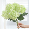 A hand holds a cluster of light green artificial hydrangea silk flowers against a white window blind.