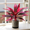 Una vibrante planta tropical rosa y morada artificial en un jarrón de cerámica gris sobre un alféizar blanco.