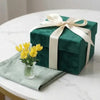 A green gift box tied with a cream ribbon sits next to a glass vase holding yellow flowers on a white marble surface.