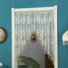 A sheer lace door curtain with light blue and green embroidery and gold tassels hanging in a white frame.