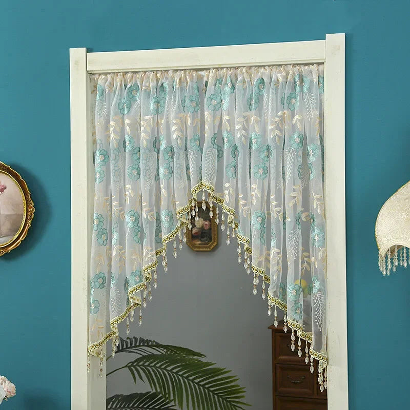 A sheer lace door curtain with light blue and green embroidery and gold tassels hanging in a white frame.