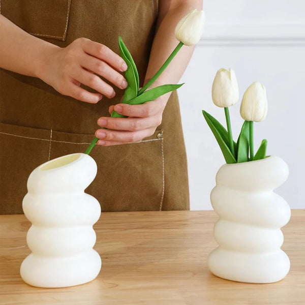 Un vase en céramique blanche à spirale tient deux tulipes blanches, tenues par une personne avec un tablier marron.