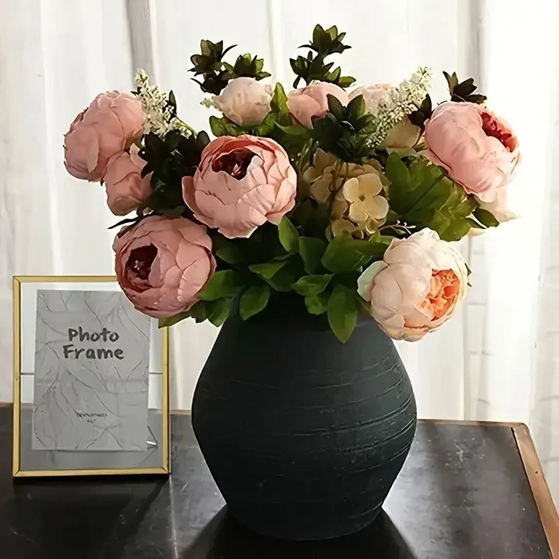 A bouquet of pink and peach peony artificial flowers in a dark vase with a photo frame.