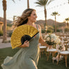 A smiling woman in a green dress holds an open gold hand fan outdoors with palm trees in the background.