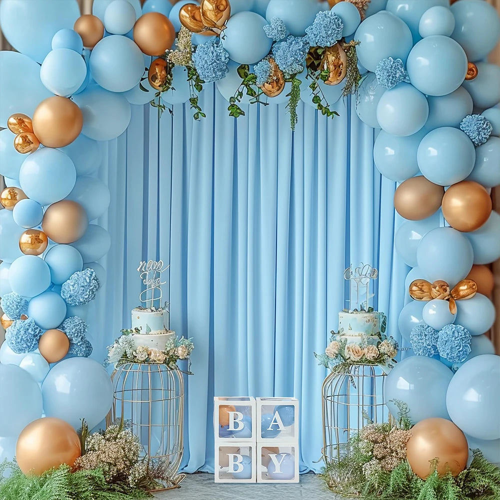 A light blue balloon and fabric backdrop decorated with gold accents and greenery for a baby shower or party.