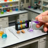 A hand holds a glass pipette with purple liquid, next to various small vials and wooden cubes on a laboratory counter.