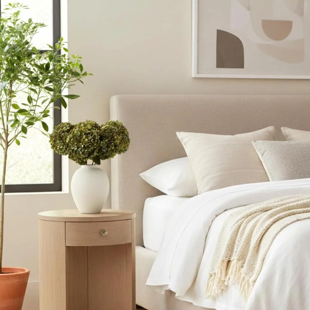 A white ceramic vase holding green foliage on a light wood nightstand next to a bed.