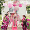 Children in pink floral dresses participate in an outdoor event surrounded by pink paper lanterns.