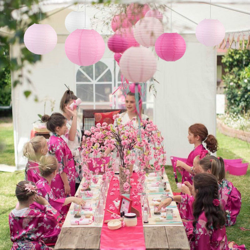 Children in pink floral dresses participate in an outdoor event surrounded by pink paper lanterns.