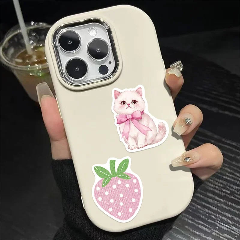 A hand holds a white phone case decorated with pink cartoon stickers of a kitten and a strawberry.