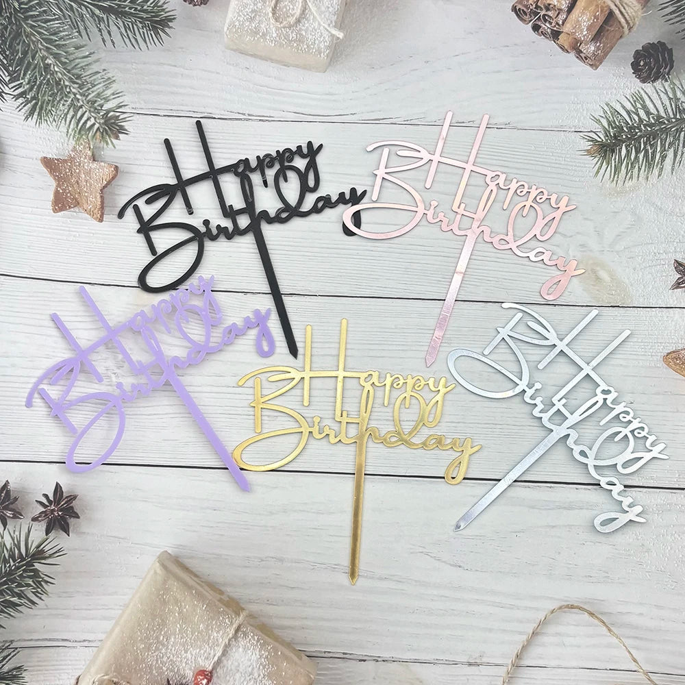 A collection of elegant acrylic birthday cake toppers with various script lettering in gold, pink, and purple on a white wood